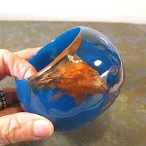 May include: A smooth, rounded blue resin bangle bracelet featuring a prominent, swirling metallic copper-colored inlay that wraps around the exterior surface, creating a high-gloss finish with a distinct contrast between the blue and copper tones.