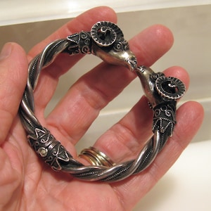 May include: A silver bracelet with two ram heads on the ends. The bracelet is twisted and has intricate designs.