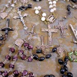 May include: A collection of rosaries with various colored beads and silver crucifixes. The rosaries are laid out on a textured surface.