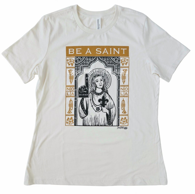 New Orleans Saints football graphic shirt in gold and black. It says Be A Saint. A saintly figure wears #1 believer foam finger, Who Dat beads, and holds a fleur de lis, with magnolias, trumpets, and NOLA border.