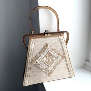 May include: Vintage ivory handbag with a decorative diamond-shaped accent. The bag features a light brown handle and frame, a gold clasp, and gold trim along the edges. The bag is made of a textured fabric.