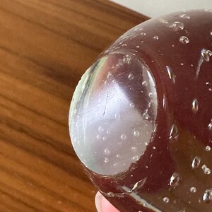 K&ouml;nnte beinhalten: Nahaufnahme eines Glasobjekts mit dunkelrotem Farbton und einem klaren, abgerundeten Bereich. Das Glas weist kleine Luftblasen auf und scheint auf einer Holzoberfl&auml;che zu liegen. Der klare Bereich hat ein paar Kratzer.