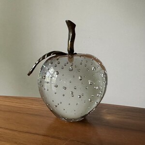K&ouml;nnte beinhalten: Eine klare Apfelskulptur aus Glas mit einem bronzefarbenen Stiel und Blatt. Der Apfel ist mit kleinen Luftblasen gef&uuml;llt, was ihm eine einzigartige Textur verleiht. Die Skulptur wird auf einer Holzoberfl&auml;che ausgestellt.