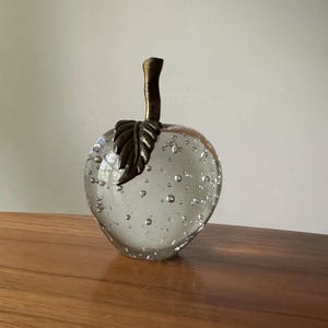 K&ouml;nnte beinhalten: Ein dekoratives Objekt in Apfelform aus klarem Glas mit Luftblasen im Inneren. Es hat einen bronzefarbenen Stiel und Blattdetails. Der Apfel steht auf einer Holzoberfl&auml;che.