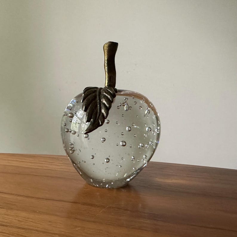 K&ouml;nnte beinhalten: Ein dekoratives Objekt in Apfelform aus klarem Glas mit Luftblasen im Inneren. Es hat einen bronzefarbenen Stiel und Blattdetails. Der Apfel steht auf einer Holzoberfl&auml;che.