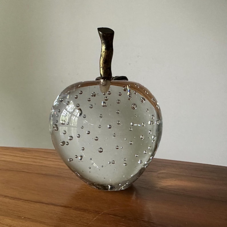 K&ouml;nnte beinhalten: Eine klare Glasapfel-Skulptur mit inneren Blasen. Der Apfel hat einen braunen Stiel mit goldenen Akzenten. Dekorative Glaskunst.