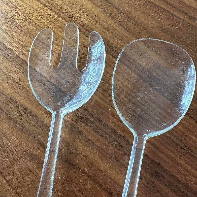 May include: A set of two clear plastic serving utensils, a fork and a spoon. The utensils are on a wooden surface.