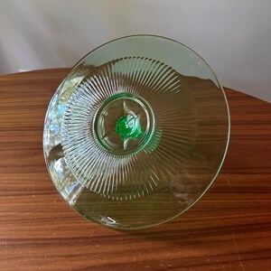 Może przedstawiać: Wazon z zielonego szkła w stylu vintage z podstawą. Naczynie ma okrągły kształt z żebrowanym wzorem promieniującym ze środka. Podstawa ma wz&oacute;r rozbłysku gwiazdy w ciemniejszym odcieniu zieleni.