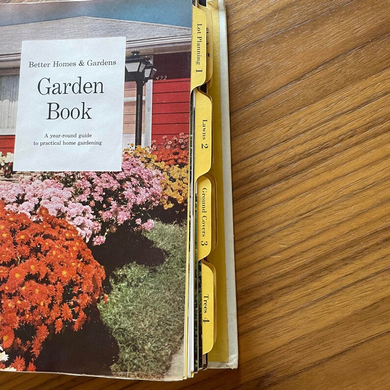 May include: A Better Homes & Gardens Garden Book with yellow index tabs labeled "Lot Planning 1", "Lawns 2", "Ground Covers 3", and "Trees 4". The book cover features a photo of a red house with a lush garden.