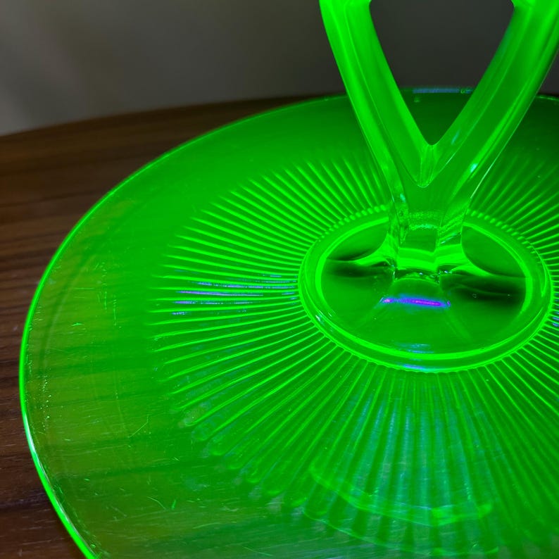 Może przedstawiać: Jasnozielony, szklany talerz do serwowania z uchwytem. Talerz ma okrągły wz&oacute;r z promienistymi liniami. Uchwyt ma kształt litery V. Szkło ma delikatny połysk, typowy dla szkła w stylu vintage. Średnica ok. 20 cm.