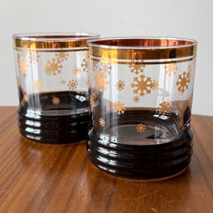 May include: Two clear drinking glasses with a gold rim and snowflake design. The base of the glasses is black. The snowflakes are gold. The glasses are sitting on a wooden surface.