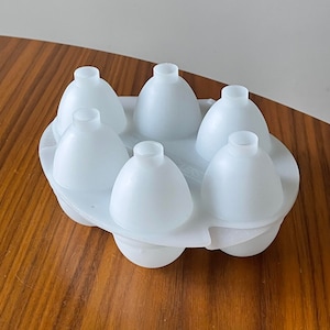 May include: A white plastic ice mold with six rounded, cone-shaped compartments. Each compartment has a small, cylindrical opening at the top. The mold is designed to create ice spheres or similar shapes for beverages.