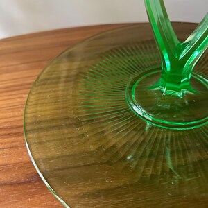 Może przedstawiać: Zielony szklany stojak na ciasto z okrągłą podstawą i n&oacute;żką. Podstawa ma żebrowany wz&oacute;r promieniujący ze środka. Stojak wykonany jest z żywego, przezroczystego zielonego szkła.