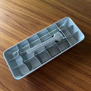 May include: A rectangular, light gray metal ice cube tray with dividers. A silver ice cube remover tool rests inside the tray. The tray is on a wooden surface. The tool has the text "General Electric" on it.