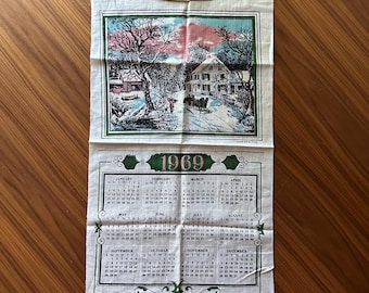 May include: A vintage 1969 calendar towel featuring a winter scene with a house, trees, and figures. The calendar grid is below the image, with months and dates in a classic layout. The towel is made of a light-colored fabric.