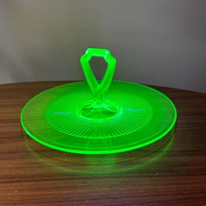 Może przedstawiać: Jasnozielony, neonowy szklany talerz do serwowania z uchwytem. Talerz jest okrągły z żebrowanym wzorem promieniującym ze środka. Uchwyt ma kształt rombu. Talerz stoi na brązowej drewnianej powierzchni.
