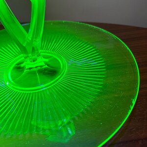 Może przedstawiać: Zbliżenie na jasną, neonową, zieloną szklaną misę do serwowania. Misa ma okrągłą podstawę z żebrowanym wzorem promieniującym ze środka. Dwa zakrzywione uchwyty rozciągają się w g&oacute;rę. Szkło ma lekki połysk.