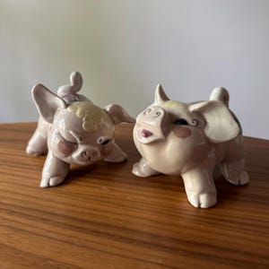 May include: Two ceramic pig figurines in a light pink hue, with detailed facial features and curled tails. One pig is winking, the other has an open mouth. The figurines are on a wooden surface.
