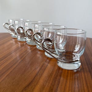 May include: A set of six clear glass teacups with silver-toned metal handles. The cups are arranged in a diagonal line on a wooden surface, showcasing their simple, elegant design. The handles are curved and provide a comfortable grip.