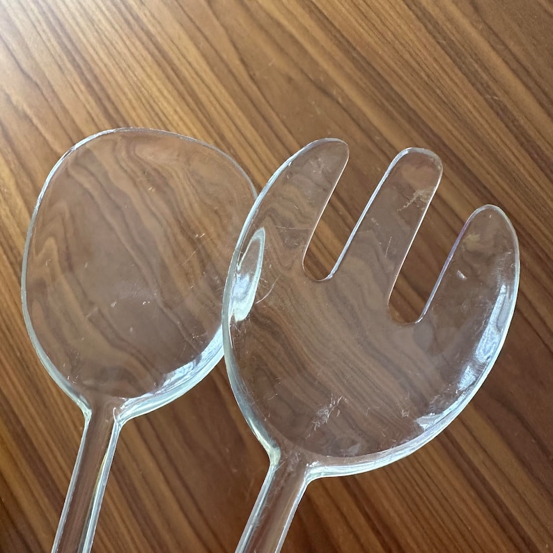May include: A set of two clear plastic salad servers. The servers are made of a clear, durable plastic and have a modern design. The larger server is a spoon shape, while the smaller server is a fork shape.