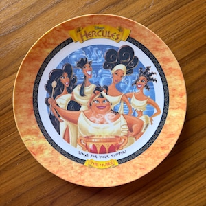 May include: Round plate featuring the Disney movie "Hercules." The plate has a light orange and brown border with a central image of the Muses. The plate has the text "Sing for your supper!" and "The Muses."