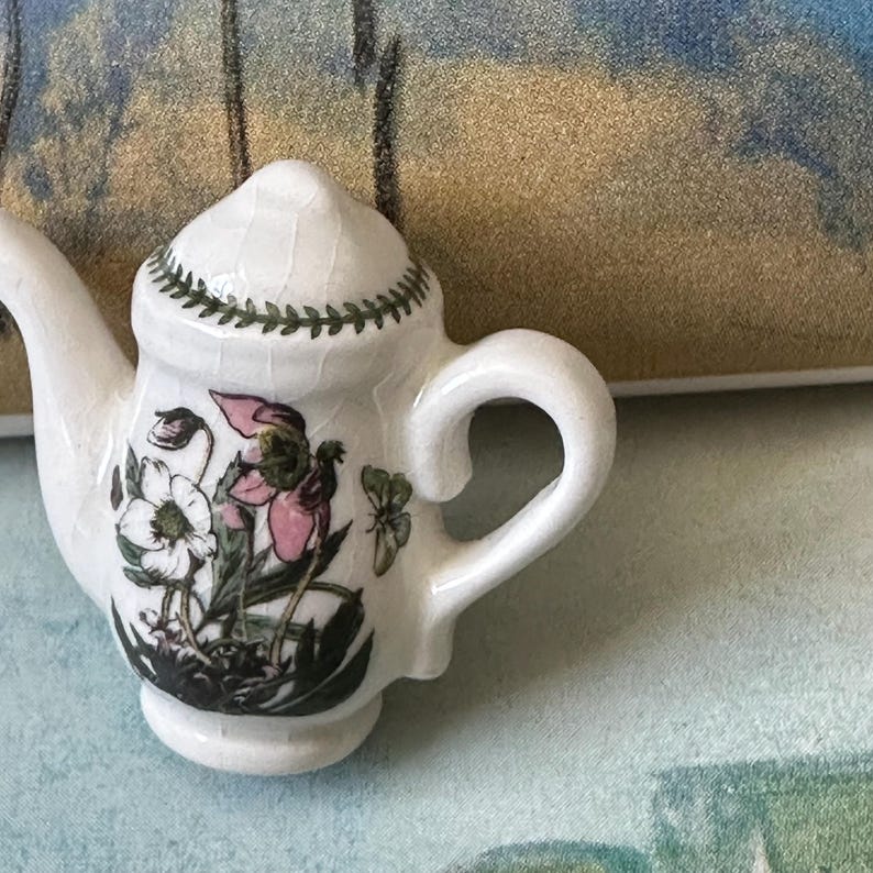 May include: A small, white ceramic teapot with a floral design featuring pink and white flowers and green leaves. The lid is decorated with a green leaf pattern. The teapot has a curved handle and a spout.