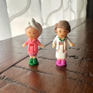 May include: Two small plastic figures. One figure wears a pink robe, green shoes, and a white towel on their head. The other figure wears a white outfit with a green collar and pink shoes.