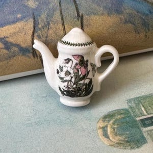 May include: A small, white ceramic teapot with a lid, spout, and handle. The teapot is decorated with a floral design in black, pink, and white. A green leaf pattern encircles the lid. The teapot is a miniature size.