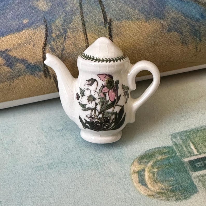 May include: A small, white ceramic teapot with a lid, spout, and handle. The teapot is decorated with a floral design in black, pink, and white. A green leaf pattern encircles the lid. The teapot is a miniature size.