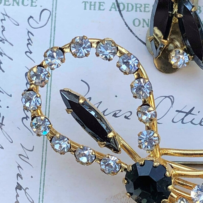 Puede incluir: Un par de pendientes de tono dorado con diamantes de imitaci&oacute;n negros y transparentes. Los pendientes tienen forma de lazo con un solo diamante de imitaci&oacute;n negro en el centro del lazo.