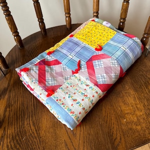 May include: A patchwork quilt made of various fabrics in shades of blue, red, yellow, and white. The quilt is folded and placed on a wooden chair. The quilt has red tassels on each corner.