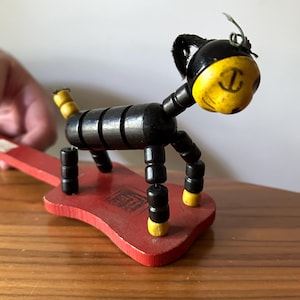 May include: A vintage wooden toy dog with a black body and yellow head, tail, and feet. The dog is mounted on a red wooden base. The toy is articulated and appears to be handmade.