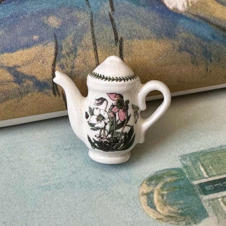 May include: A small, white ceramic teapot with a floral design featuring pink and white flowers and green leaves. The teapot has a curved handle, a spout, and a lid with a green border. The teapot is a miniature size.