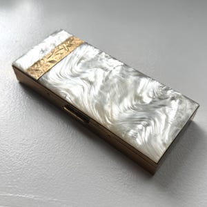 May include: A rectangular metal case featuring a white pearlescent marbled top panel. A gold-toned metal strip with an engraved floral pattern accents one end of the lid. The sides of the case are finished in a smooth, solid gold-toned metal.