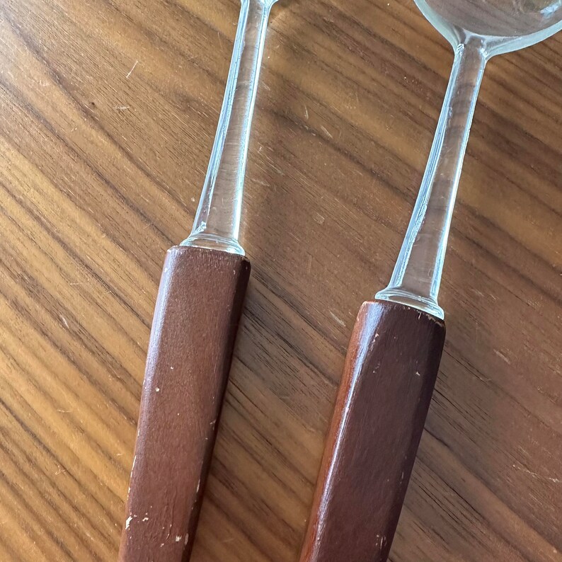 May include: A set of two wooden handled serving spoons with clear glass bowls. The spoons are made of dark brown wood and have a smooth finish.