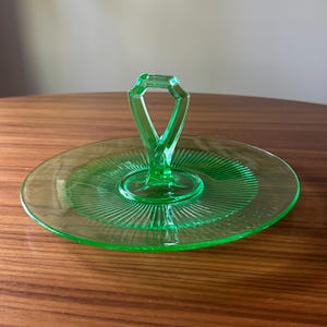 Może przedstawiać: Zabytkowa zielona szklana taca do serwowania z centralnym uchwytem. Okrągła taca ma żebrowany wz&oacute;r promieniujący ze środka. Uchwyt ma geometryczny kształt. Taca jest prezentowana na drewnianej powierzchni.
