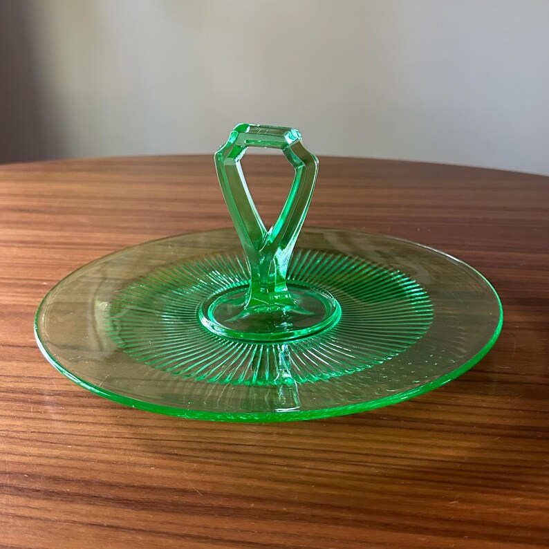 Może przedstawiać: Zabytkowa zielona szklana taca do serwowania z centralnym uchwytem. Okrągła taca ma żebrowany wz&oacute;r promieniujący ze środka. Uchwyt ma geometryczny kształt. Taca jest prezentowana na drewnianej powierzchni.