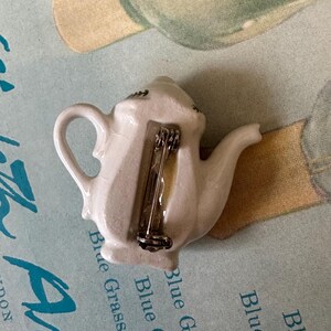 May include: White ceramic teapot pin with a silver pin back. The teapot has a curved handle and spout, with a small decorative design on the top. The pin is likely a vintage or collectible item, perfect for tea lovers or those who enjoy unique accessories.