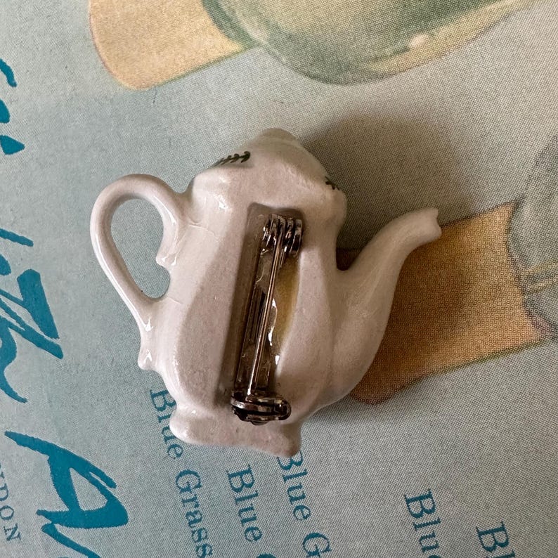 May include: White ceramic teapot pin with a silver pin back. The teapot has a curved handle and spout, with a small decorative design on the top. The pin is likely a vintage or collectible item, perfect for tea lovers or those who enjoy unique accessories.