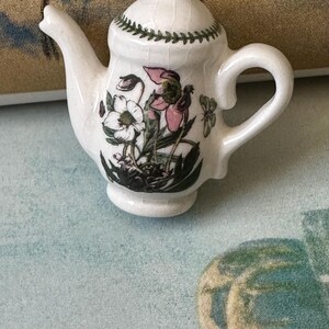 May include: A small, white ceramic teapot with a floral design featuring pink and white flowers and green leaves. The teapot has a curved handle, a spout, and a lid with a green leaf pattern around the edge. The teapot is a miniature size.