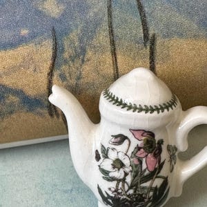 May include: A small, white ceramic teapot with a floral design featuring pink and white flowers and green leaves. The lid is decorated with a green leaf pattern. The teapot has a curved spout and handle.
