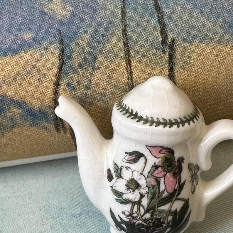 May include: A small, white ceramic teapot with a floral design featuring pink and white flowers and green leaves. The lid is decorated with a green leaf pattern. The teapot has a curved spout and handle.