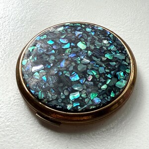 May include: A round compact mirror with a brass-toned metal frame. The lid features an inlay of crushed abalone shell fragments in shades of iridescent blue, green, and purple, creating a mosaic pattern across the entire circular surface area.