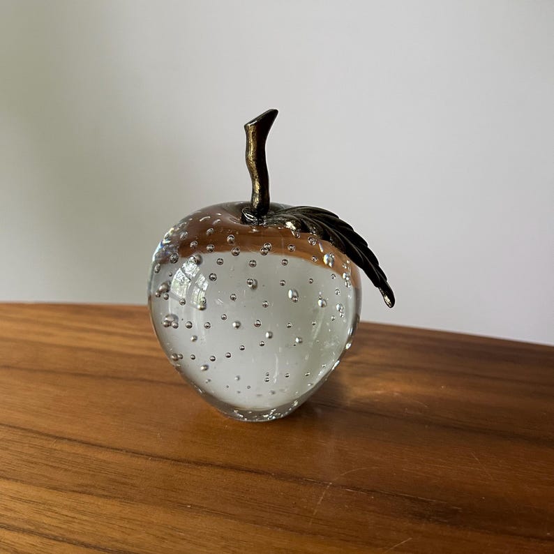 K&ouml;nnte beinhalten: Ein dekorativer Apfel aus klarem Glas mit einem Bronzestiel und Blatt. Der Apfel hat ein wei&szlig;es Inneres und ist mit kleinen Luftblasen gef&uuml;llt. Die Oberseite des Apfels ist hellbraun. Das Objekt steht auf einer Holzoberfl&auml;che.