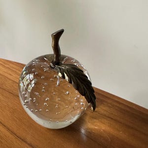 K&ouml;nnte beinhalten: Ein klarer Glas-Apfel-Briefbeschwerer mit einem Bronzestiel und Blatt. Das Glas ist mit kleinen Blasen gef&uuml;llt, und der Boden ist mattwei&szlig;. Der Apfel liegt auf einer Holzoberfl&auml;che.