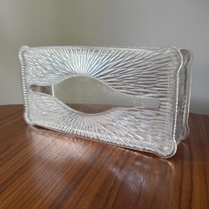 May include: A rectangular clear glass tissue box cover featuring a pressed sunburst pattern radiating from the center oval opening. The edges of the glass container are slightly curved with a decorative molded texture throughout the entire surface.