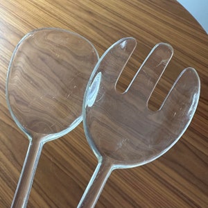 May include: A set of two clear plastic salad servers. The server on the left is a spoon shape and the server on the right has a fork shape.