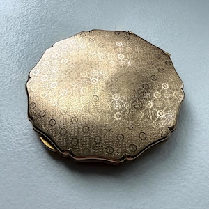 May include: A round, gold-toned metal compact case featuring a scalloped edge and an intricate, repeating floral pattern engraved across the entire top surface. The metallic finish has a reflective quality, highlighting the detailed texture of the design.