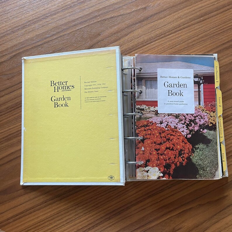 May include: A yellow Better Homes and Gardens Garden Book with a revised edition copyright 1961, 1971, 1981. The book is open to a page with a photo of a red brick house with a flower garden in front. The text on the page reads "Better Homes & Gardens Garden Book A year-round guide to practical home gardening."