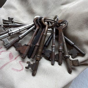 May include: A collection of antique keys on a metal ring. The keys vary in size and shape, with a mix of silver and rusted brown finishes. The keys are arranged on a beige fabric background.